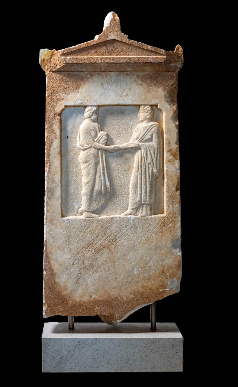 11. Μαρμάρινη αττική επιτύμβια στήλη με αετωματική επίστεψη. Φέρει ανάγλυφη παράσταση σε ορθογώνιο διάχωρο. Απεικονίζεται σκηνή δεξίωσης ανάμεσα σε ανδρική και γυναικεία μορφή. Ύψος 90,2 εκ. Αρχές 4ου αι. π.Χ.